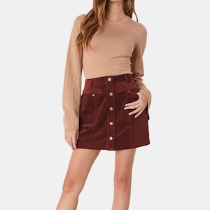 Può includere: Una donna indossa un maglione color cammello a maniche lunghe e una minigonna in velluto a coste bordeaux. La gonna ha una chiusura con bottoni e tasche. L'outfit è casual, a tema autunnale.