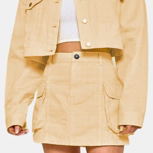 May include: A light tan denim jacket and matching mini skirt set. The cropped jacket has button closures and chest pockets. The skirt features cargo pockets and a button closure. The outfit is a solid color.