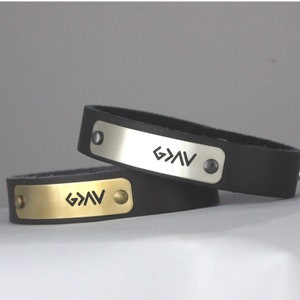May include: Two leather bracelets with metal plates engraved with the symbol "G>>V". One bracelet has a silver plate and the other has a gold plate.