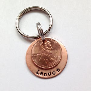 May include: A copper keychain with a penny attached to it. The penny is dated 2014 and has the words "In God We Trust" and "Liberty" on it. The keychain has the name "Landon" engraved on it.