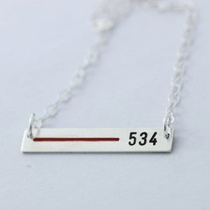 May include: A silver bar necklace with a red line and the number 534 in black. The necklace has a delicate chain and a rectangular pendant. The background is a soft, neutral color.