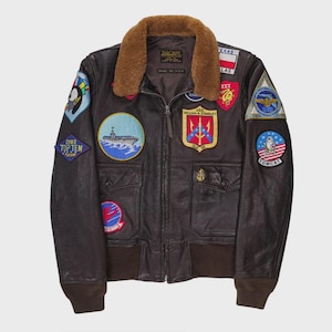 May include: A brown leather bomber jacket with a shearling collar and various embroidered patches. The jacket has a zip-up front, two front pockets, and ribbed cuffs and hem. Patches include military insignias and the word "Texas".