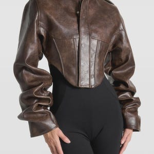 May include: A brown leather jacket with a high collar and a fitted waist. The jacket has long sleeves with a gathered design at the wrists. The jacket is paired with black pants.