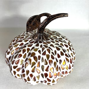 May include: A decorative pumpkin with a mosaic design. The pumpkin is white with brown, gold, and amber-colored glass pieces. It has a bronze-colored stem and leaf. A fall-themed decorative item.