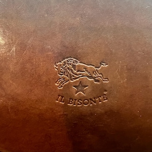 May include: Close-up of a brown leather item with the embossed logo of a lion over a star and the words "IL BISONTE". The leather has a textured appearance with variations in color.