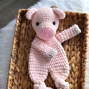 May include: A handmade, light pink crocheted pig plush toy with black eyes, a pink snout, and gray accents on its feet and hands. The pig is lying in a woven basket, creating a cozy and inviting scene.