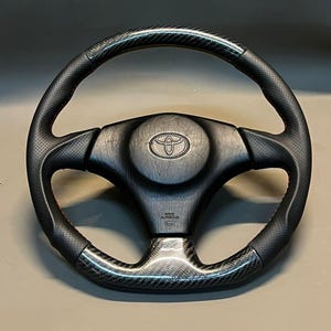 Könnte beinhalten: Schwarzes Lenkrad mit Carbonfaser- und perforiertem Lederdesign. Das Lenkrad hat rote Nähte und ein Toyota-Logo in der Mitte. Das Wort "AIRBAG" ist sichtbar.