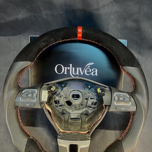 Puede incluir: Un volante de cuero negro y gris perforado con costuras rojas y una franja roja en la parte superior. El volante tiene controles integrados y la marca "Orluvéa" es visible en el fondo.