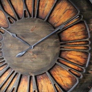 Può includere: Un grande orologio rotondo in legno con una cornice marrone scuro e un centro marrone più chiaro. Il quadrante presenta numeri romani e lancette metalliche. Gli elementi in legno hanno un aspetto invecchiato e rustico.