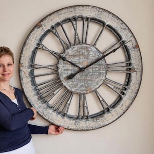 Pode incluir: Um relógio de parede grande e redondo com um acabamento desgastado em branco e cinzento. O relógio apresenta um design de estrutura aberta com algarismos romanos pretos proeminentes e ponteiros de metal preto finos. A superfície mostra desgaste.