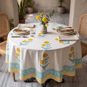 May include: A round white tablecloth with a yellow and blue floral pattern. The tablecloth is set on a round table with a black base. The table is set with plates, silverware, and glasses. A vase of yellow flowers sits in the center.