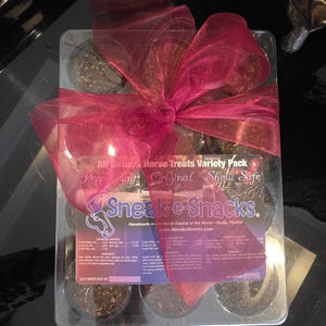 May include: A clear plastic container holding a variety pack of horse treats, topped with a large magenta ribbon. The label reads Sneak-e-Snacks, All Natural Horse Treats Variety Pack, Peppermint, Original, and Show Safe, with listed ingredients.
