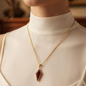 Könnte beinhalten: Eine goldfarbene Halskette mit einem roten, rautenförmigen Anhänger. Der Anhänger ist in Gold gefasst und hängt an einer feinen Kette. Die Halskette wird über einem weißen Rollkragen getragen.