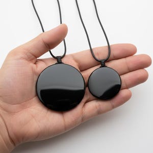 Może przedstawiać: Dwa czarne, okrągłe wisiorki na czarnych sznurkach. Jeden wisiorek jest większy od drugiego. Wisiorki są gładkie i błyszczące, odbijają światło. Sznurki są zawiązane na górze. Wisiorki są trzymane w dłoni na białym tle.