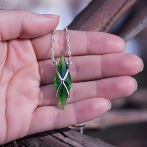 May include: A silver necklace featuring a green, gem-like pendant. The pendant has a pointed, crystal-like shape with a silver metal cage around it. The necklace is held in a hand, with a blurred background.