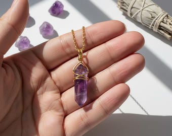 Pode incluir: Um colar de corrente dourada com um pingente de cristal de ametista roxa. O cristal é envolto em fio de ouro. Várias pedras pequenas de ametista estão espalhadas ao redor do colar. Um maço de sálvia está ao fundo.