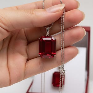 Puede incluir: Un collar de plata con un gran colgante rectangular de piedra preciosa rojo intenso. El colgante está engastado en un marco de plata y cuelga de una delicada cadena de plata. Un colgante a juego es visible en el fondo.