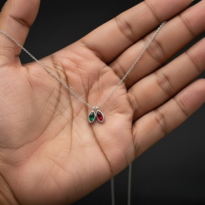 Pode incluir: Um colar de prata com uma corrente delicada. O pingente apresenta duas pedras preciosas em corte marquise, uma verde esmeralda e outra vermelho rubi, lado a lado. O colar é exibido em um fundo escuro.