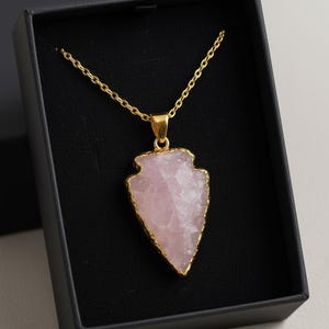 May include: A gold-colored chain necklace with a pendant shaped like an arrowhead. The pendant is a light pink stone with gold-colored edging, presented in a black jewelry box.