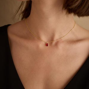 May include: A delicate gold necklace featuring a small, teardrop-shaped red gemstone pendant. The necklace is worn against a black top, highlighting the contrast between the gold chain and the vibrant red stone.