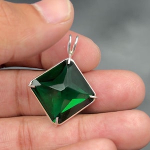 Puede incluir: Un colgante cuadrado de piedra preciosa verde esmeralda con una fianza plateada. La piedra preciosa está facetada, captando la luz y mostrando varios tonos de verde. El colgante se sostiene en una mano sobre un fondo gris.