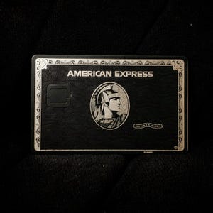 Puede incluir: Una tarjeta de crédito American Express negra con detalles plateados. La tarjeta muestra "AMERICAN EXPRESS" en la parte superior y un perfil de centurión romano. La tarjeta tiene una superficie texturizada y una ranura para chip.