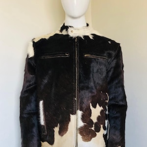 May include: A black and white cowhide jacket with a zip-up front and two chest pockets. The jacket has a stand-up collar and a brown leather trim. The jacket is displayed on a mannequin.