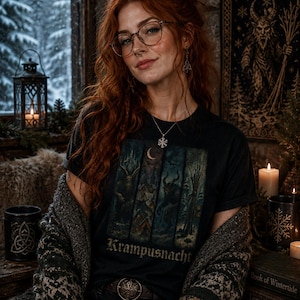 May include: A black t-shirt featuring a graphic print with four vertical panels depicting winter scenes, a crescent moon, and the text Krampusnacht. The shirt is worn with a dark green patterned cardigan, a silver snowflake necklace, and glasses.