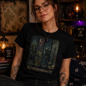 May include: A black t-shirt featuring a graphic print divided into four vertical panels depicting a dark winter scene with a horned figure, a crescent moon, and a snowy village. The text Krampusnacht is printed in a gothic font at the bottom.