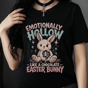 Puede incluir: Camiseta negra con el texto "Emotionally Hollow - Like a Chocolate Easter Bunny". El gráfico presenta un conejito con detalles esqueléticos expuestos. El diseño incluye colores azul pastel y rosa.