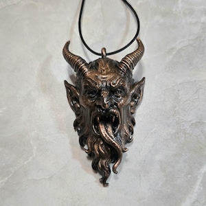 May include: A bronze-colored pendant necklace featuring a detailed, demonic face with horns, fangs, and a long tongue. The pendant is suspended from a black cord. The design has a gothic or fantasy theme.