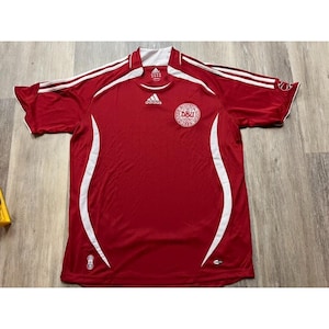 May include: A red short-sleeve jersey featuring white side panels and white stripes along the shoulders. The Adidas logo is centered on the chest, with the DBU crest on the left side. A small emblem is visible on the lower left front hem.