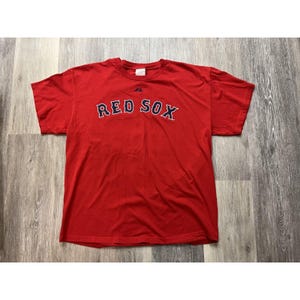 Può includere: Una maglietta rossa con la scritta "RED SOX" in lettere blu navy e bianche. La maglietta ha maniche corte e girocollo. Il logo del marchio Majestic è visibile sul petto.