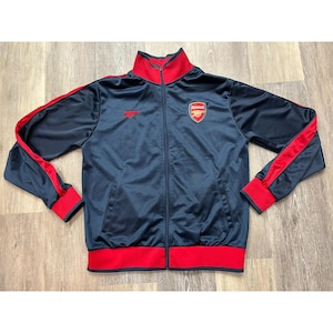 May include: Navy blue track jacket with red detailing. Includes a red collar, cuffs, and stripes along the sleeves. The jacket has a full zip and the Arsenal logo on the chest.