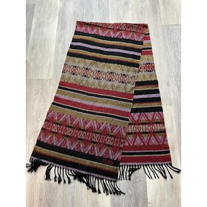 May include: A Fendi scarf with a geometric pattern in red, black, gold, and purple. The scarf has a fringed edge and a woven design. The scarf is made of a soft material and is suitable for cold weather.