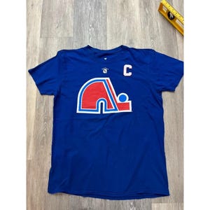 Puede incluir: Una camiseta azul real con un gran logotipo con detalles en rojo, blanco y azul. El logotipo es un diseño estilizado con una "C" blanca en la parte superior derecha. La camiseta es de manga corta.