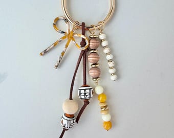 May include: A gold-colored keychain with a large round clasp. The keychain features a decorative bow and several strands of beads in various colors, including white, brown, and yellow. Some beads have geometric patterns.