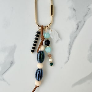 May include: Gold-coloured carabiner keyring with decorative beads in black, blue, white, and green. A leather accent and a white, leaf-shaped charm are also attached. The keyring is designed for keys or accessories.