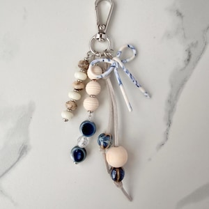 May include: A decorative keychain with a silver clasp, featuring a variety of beads in cream, brown, and blue hues. The design includes wooden beads, ceramic beads, and a small bow with blue and white patterns. A stylish accessory.