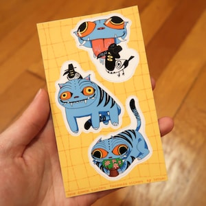 May include: A rectangular yellow sticker sheet with a grid pattern featuring three blue creature stickers. One shows a blue head with a long tongue, another a blue cat-like creature with a small bird, and the third a blue cat holding a potted plant.