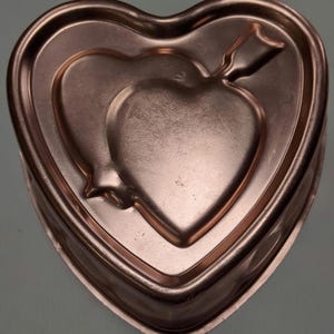 Może przedstawiać: Miedziana forma do pieczenia w kształcie serca z motywem serca pośrodku. Forma ma podniesioną krawędź i dekoracyjny wzór strzały. Forma jest wykonana z metalu i ma błyszczące wykończenie.