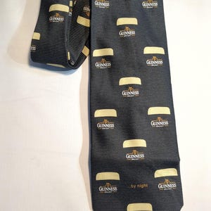 May include: Black tie featuring a repeating pattern of Guinness beer glasses and the Guinness logo in gold. The tie is made of a ribbed fabric and includes the text "...by night". A stylish accessory for any occasion.