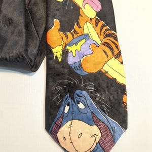 Puede incluir: Corbata negra con Tigger y Eeyore de Winnie the Pooh. Tigger es naranja y negro, sosteniendo un tarro de miel. Eeyore es azul con una expresión triste. La corbata tiene un patrón de espiga negro.