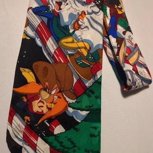 Puede incluir: Una corbata de seda colorida con un diseño navideño con Bugs Bunny, Yosemite Sam y Piolín. La corbata presenta un fondo negro con bastones de caramelo rojo y blanco, árboles de Navidad verdes y nieve blanca.