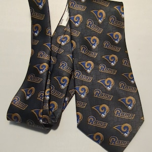 May include: A black necktie featuring a repeating pattern of the Los Angeles Rams logo and the word "Rams" in gold and blue. The tie has a textured background and is made of a smooth fabric.
