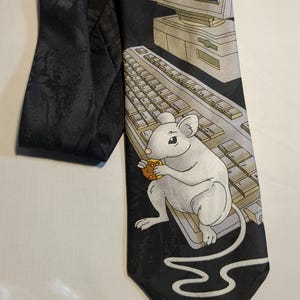 Könnte beinhalten: Schwarze Krawatte mit einem Computer-Motiv. Die Krawatte zeigt eine weiße Maus, die einen Keks hält, eine Tastatur, einen Computermonitor und einen Computer-Tower. Der Hintergrund ist schwarz.