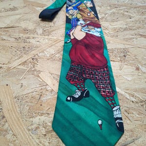 May include: A green, black, and red silk tie with a cartoon image of Santa Claus and other golfers playing on a green golf course.