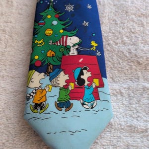 May include: A blue necktie featuring a Peanuts holiday scene with Snoopy and Woodstock near a decorated Christmas tree and red doghouse. Below, three characters in winter clothing are singing in the snow, which fades to a light blue color.