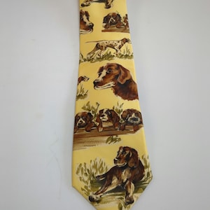 Puede incluir: Una corbata de seda amarilla con un patrón repetido de perros marrones y blancos. Los perros están en varias poses, incluyendo sentados, de pie y corriendo. La corbata tiene una punta puntiaguda.