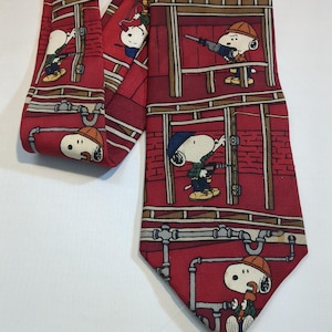 Puede incluir: Corbata roja con un patrón repetido de Snoopy como trabajador de la construcción. La corbata presenta un diseño de pared de ladrillo y tuberías. Snoopy lleva un casco y usa herramientas.